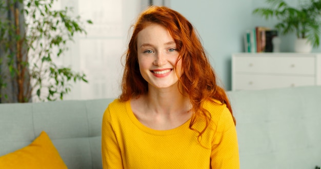 Ritratto di bella ragazza allegra di redhead che sorride esaminando macchina fotografica che si siede sul sofà nel paese