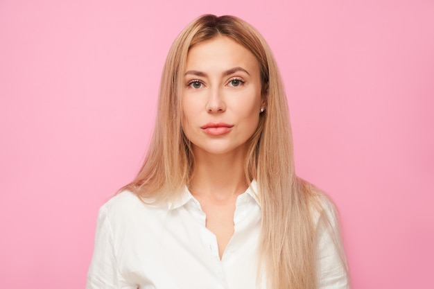 Ritratto di bella ragazza bionda dell'ufficio in camicia bianca che esamina la macchina fotografica isolata sul fondo rosa dello studio