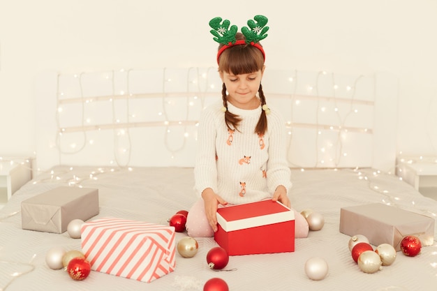 Ritratto di bella ragazza carina che indossa un maglione bianco e corna di cervo carnevale cerchio apertura scatola regalo di Natale con interesse, festa di Capodanno, vacanze invernali.