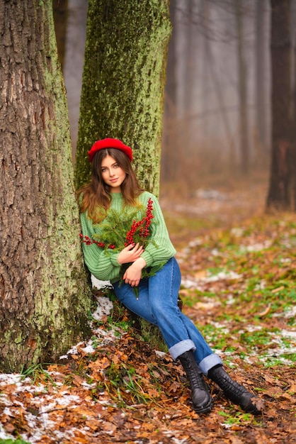 Ritratto di bella ragazza castana in cappello rosso e maglione lavorato a maglia verde che tiene i rami di Natale