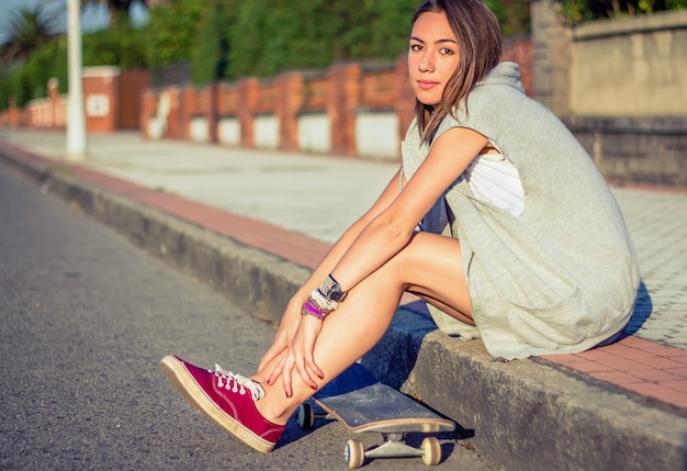 Ritratto di bella ragazza con pantaloncini corti e skateboard seduti all'aperto in una calda giornata estiva. Edizione dai toni caldi.