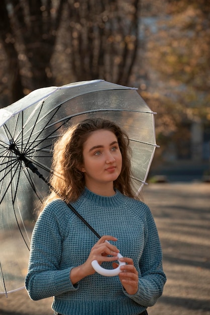 Ritratto di bella ragazza pensierosa con occhi azzurri e capelli ricci sotto l'ombrello trasparente Giovane donna in maglione blu