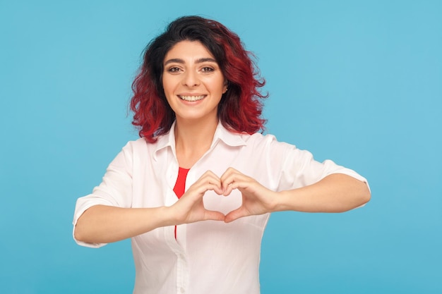 Ritratto di bella romantica donna felice con fantasia capelli rossi che fanno a forma di cuore con le mani e sorride alla telecamera gesticolando amore speranza carità segno indoor studio girato isolato su sfondo blu