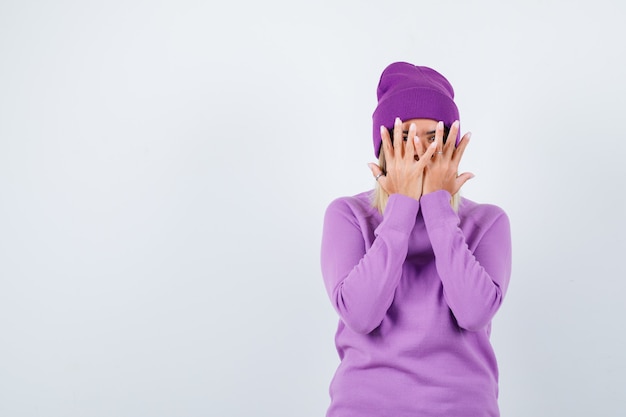 Ritratto di bella signora che copre il viso con le mani, guardando attraverso le dita in maglione, berretto e guardando la vista frontale spaventata