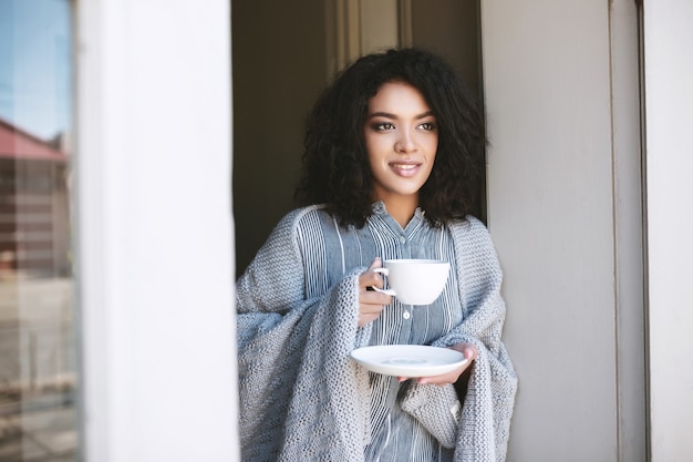 Ritratto di bella signora con capelli ricci scuri che si appoggia sulla porta con una tazza di caffè in mano