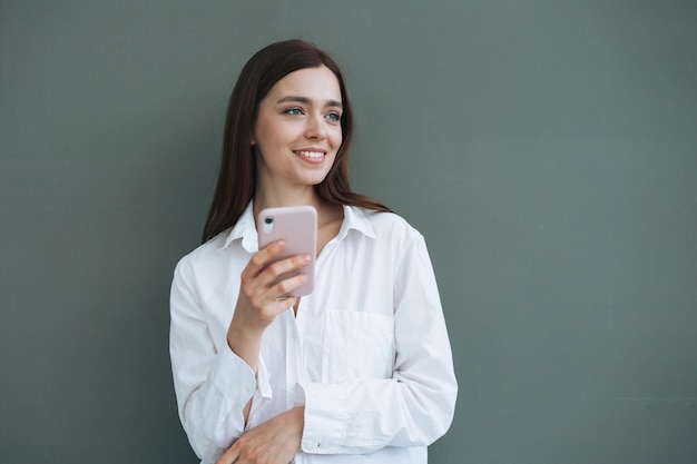 Ritratto di bella studentessa sorridente con i capelli lunghi scuri in camicia bianca utilizzando il telefono cellulare in mano su sfondo grigio isolato generazione Z