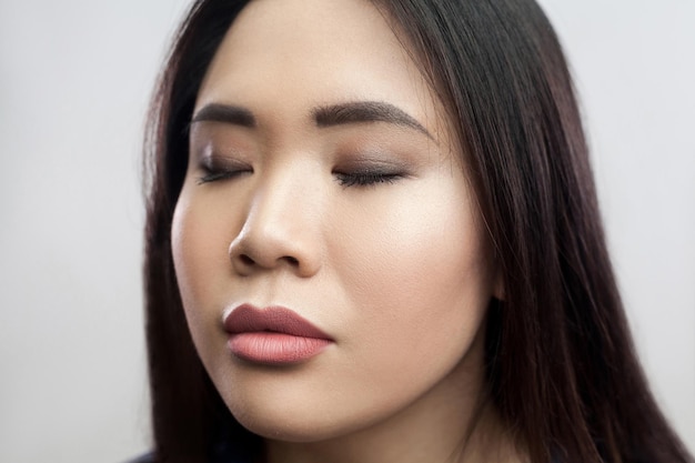 Ritratto di bellezza del primo piano della giovane donna asiatica castana bella calma con il trucco, capelli scuri lisci che stanno con gli occhi chiusi e la faccia rilassata. ripresa in studio indoor, isolata su sfondo grigio chiaro.