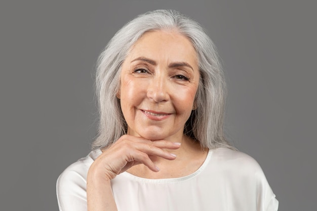 Ritratto di bellezza della donna anziana che tocca il viso guardando la fotocamera e sorridente su studio grigio