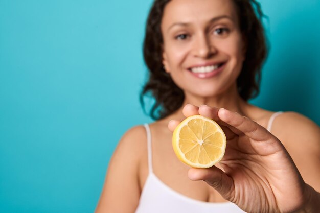 Ritratto di bellezza della giovane donna sorridente allegra attraente con i capelli scuri ricci in cima bianca isolata su fondo blu luminoso che tiene metà del limone e che copre gli occhi con esso. Copia spazio pubblicitario