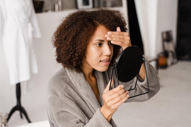 Ritratto di bellezza della ragazza afroamericana in accappatoio La donna attraente sta guardando l'acne e i brufoli sul viso Routine di bellezza mattutina per la cura della pelle in bagno