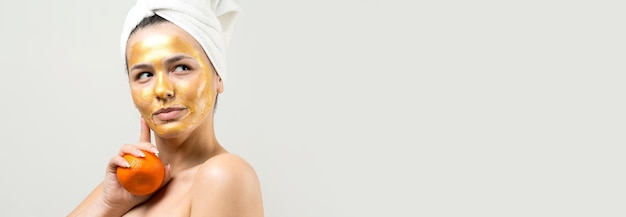 Ritratto di bellezza di donna in asciugamano bianco sulla testa con maschera nutriente d'oro sul viso Pulizia della pelle eco cosmetici biologici spa relax concept Una ragazza sta con la schiena tenendo un mandarino arancione