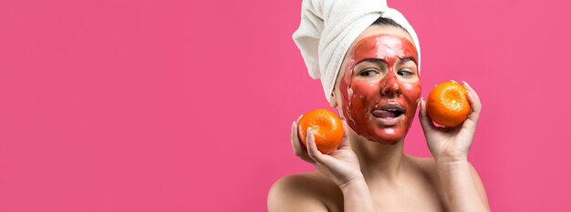 Ritratto di bellezza di donna in asciugamano bianco sulla testa con maschera nutriente rossa sul viso Pulizia della pelle eco cosmetici biologici spa relax concept Una ragazza sta con la schiena tenendo un mandarino arancione