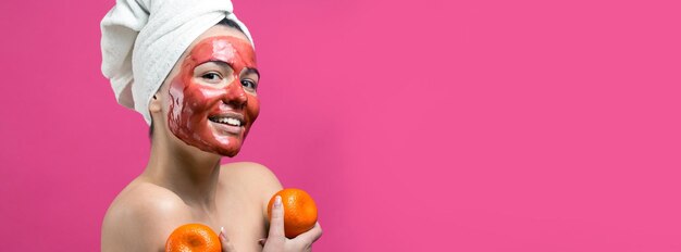 Ritratto di bellezza di donna in asciugamano bianco sulla testa con maschera nutriente rossa sul viso Pulizia della pelle eco cosmetici biologici spa relax concept Una ragazza sta con la schiena tenendo un mandarino arancione