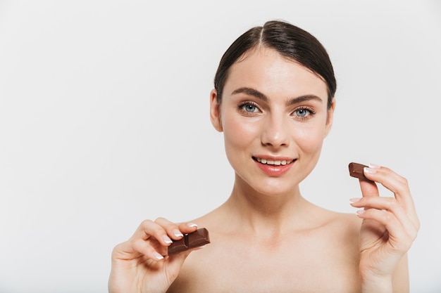 Ritratto di bellezza di un'attraente giovane donna bruna in piedi isolata sul muro bianco, che mostra una barretta di cioccolato nero