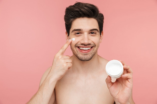 Ritratto di bellezza di un bell'uomo bruna senza camicia sorridente in piedi isolato su un muro rosa, applicando la crema per il viso da un contenitore