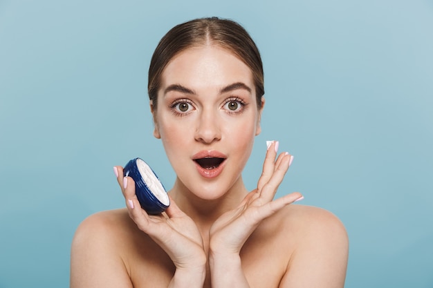 Ritratto di bellezza di una giovane donna attraente in topless isolata sul muro blu, applicando la crema da un contenitore