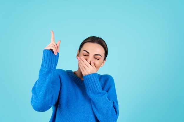 Ritratto di bellezza di una giovane donna con un maglione lavorato a maglia blu e un trucco da giorno naturale, che sorride allegramente e punta il dito verso uno spazio vuoto