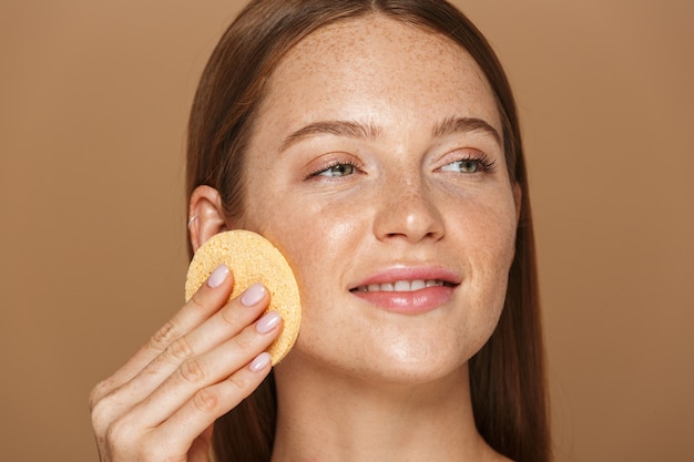 Ritratto di bellezza di una giovane donna sorridente in topless con lunghi capelli rossi che pulisce il viso con una spugna isolata sopra il muro beige