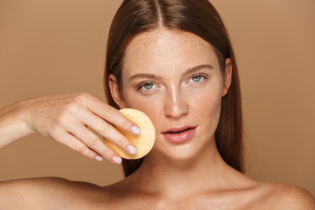 Ritratto di bellezza di una giovane donna sorridente in topless con lunghi capelli rossi che pulisce il viso con una spugna isolata sopra il muro beige