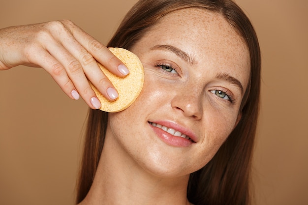 Ritratto di bellezza di una giovane donna sorridente in topless con lunghi capelli rossi che pulisce il viso con una spugna isolata sopra il muro beige