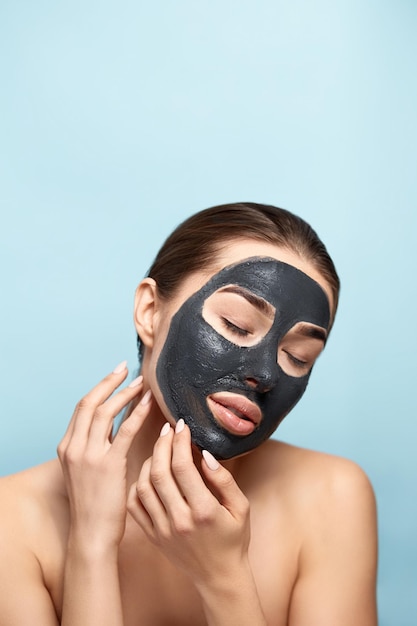 Ritratto di bellezza donna cura della pelle salute maschera nera sfondo blu primo piano Modello ragazza tocca il viso Maschera cosmetica Trattamento termale Cosmetologia