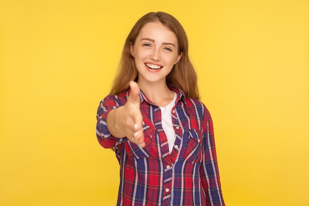 Ritratto di benvenuto di una ragazza di zenzero socievole e amichevole in camicia a scacchi che allunga la mano alla telecamera offrendo un saluto di stretta di mano e facendo conoscenza con uno studio al coperto isolato su sfondo giallo