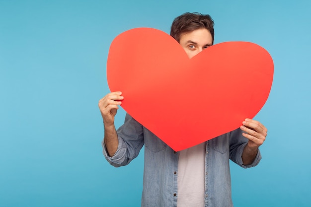 Ritratto di bruna uomo giocoso in camicia di jeans che si nasconde dietro il cuore di carta rossa che sbircia fuori dall'enorme biglietto di auguri di San Valentino che dimostra l'amore in studio indoor girato isolato su sfondo blu