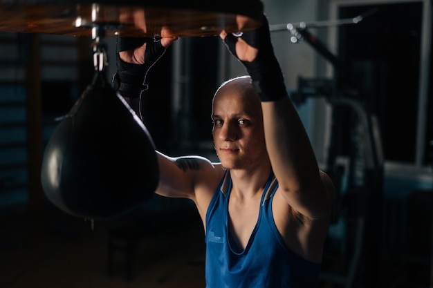 Ritratto di brutale pugile calvo maschio in nastro di difesa della boxe che riposa vicino al sacco da boxe in un club sportivo con interni scuri, guardando la fotocamera. Combattente stanco che si rilassa dopo l'allenamento.