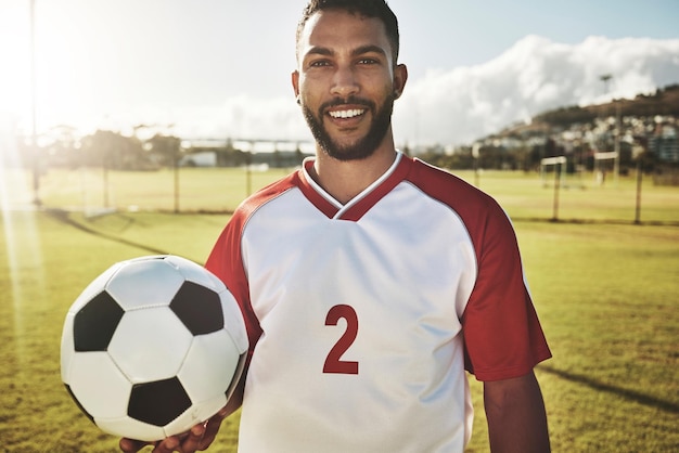 Ritratto di calciatore e uomo con un pallone da calcio in un campo sorriso felice ed entusiasta dell'obiettivo sportivo nel campo di calcio Giocatore di calcio fitness e ragazzo in un campo di calcio per l'allenamento