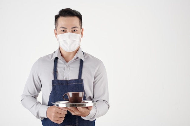 Ritratto di cameriere di coffeeshop in maschera medica che serve tazza di caffè appena fatto