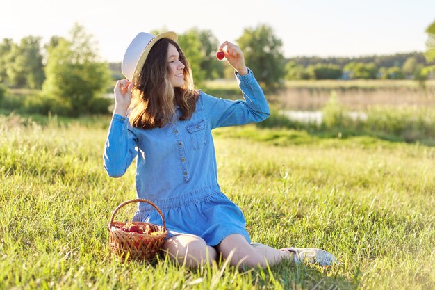 Ritratto di campagna di bella ragazza adolescente in cappello di jeans con cesto di fragole fresche, mangiando fragole mature. Sfondo del paesaggio naturale, prato verde, cielo blu