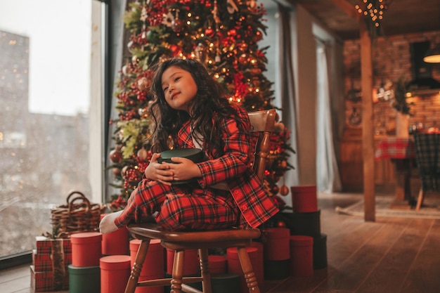 Ritratto di candida bambina sorridente asiatica in pigiama plaid rosso seduto con regali a casa di Natale