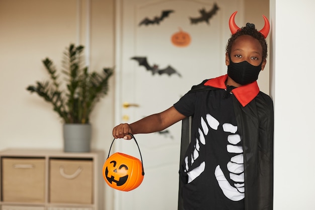 Ritratto di carino ragazzo afroamericano che indossa un costume da diavolo spettrale con corna rosse e maschera che tiene jack o 'lanterna cestino per caramelle