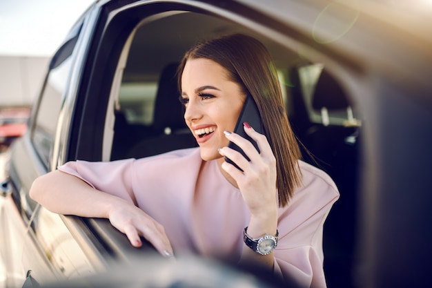 Ritratto di castana elegantemente vestito splendido che per mezzo dello Smart Phone mentre appoggiandosi il finestrino della macchina.