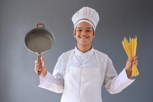 Ritratto di chef sorridente in toque bianco e grembiule che tiene padella e spaghetti isolati su gre