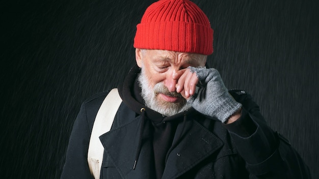 Ritratto di Close-up di un uomo di 70 anni in pensione con una faccia rugosa, vestito con un cappotto e un cappello rosso, asciuga le lacrime dagli occhi su uno sfondo nero isolato