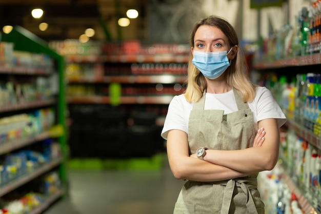 Ritratto di commessa, in maschera protettiva donna sorridente e guardando la fotocamera in un supermercato. Venditore femminile amichevole piacevole che sta nel deposito fra le file. operaio con le braccia incrociate