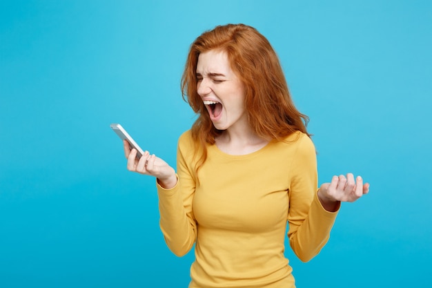 Ritratto di concetto di tecnologia e stile di vita della ragazza rossa dei capelli dello zenzero che grida sul telefono isolato sullo spazio pastello blu della copia della parete