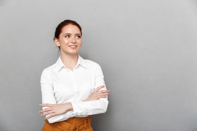 Ritratto di contenuto redhead imprenditrice 20s in abbigliamento formale sorridente e in posa in ufficio isolato su gray
