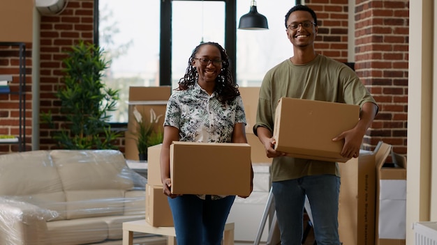 Ritratto di coppia afroamericana che si trasferisce insieme dopo l'acquisto di un appartamento in prestito ipotecario. Guardare la telecamera e sentirsi felici per il trasloco, contenitori di cartone per spostare i mobili.