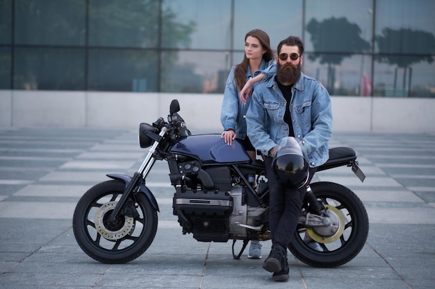 Ritratto di coppia attraente in giacche di jeans con moto vicino al grande edificio di vetro nel centro della città.