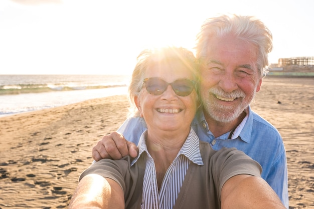 Ritratto di coppia di persone mature e anziane che si godono l'estate in spiaggia guardando la telecamera prendendo un selfie insieme al tramonto sullo sfondo. Due anziani attivi che viaggiano all'aperto.