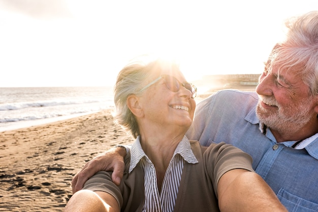 Ritratto di coppia di persone mature e anziane che si godono l'estate in spiaggia guardando la telecamera prendendo un selfie insieme al tramonto sullo sfondo. Due anziani attivi che viaggiano all'aperto.