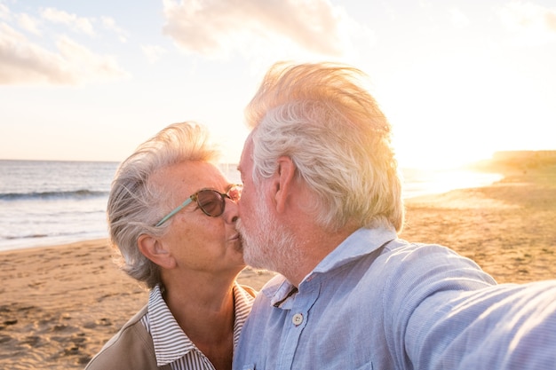 Ritratto di coppia di persone mature e anziane che si godono l'estate in spiaggia guardando la telecamera prendendo un selfie insieme al tramonto sullo sfondo. Due anziani attivi che viaggiano all'aperto.