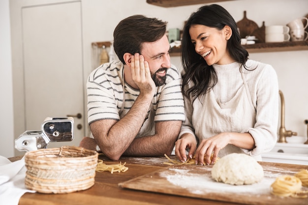 Ritratto di coppia positiva uomo e donna 30s che indossano grembiuli che fanno pasta fatta in casa di pasta mentre cucinano insieme in cucina a casa