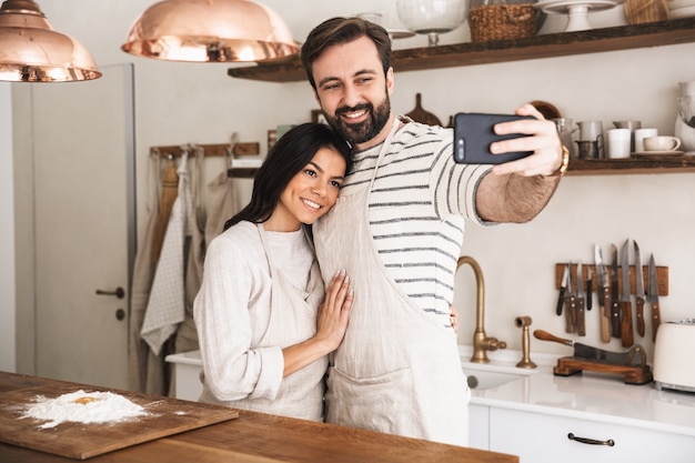 Ritratto di coppia positiva uomo e donna 30s che indossano grembiuli che scattano foto selfie mentre cucinano la pasticceria con farina e uova in cucina a casa