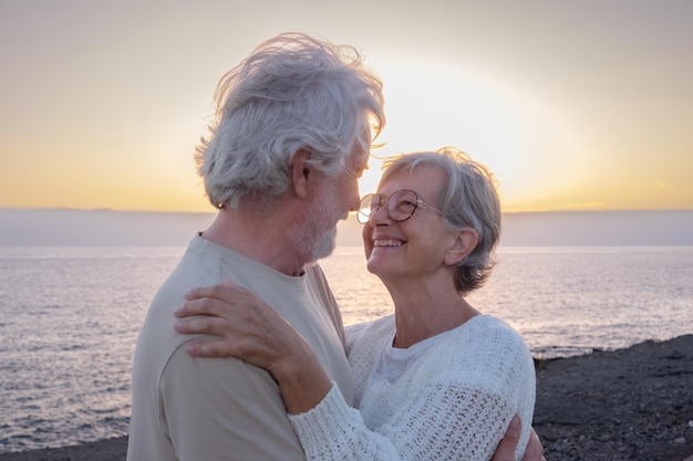 Ritratto di coppia senior felice e romantica o in pensione abbracciata al mare alla luce del tramonto che si guarda negli occhi vecchia coppia senior sorridente all'aperto che si gode le vacanze insieme