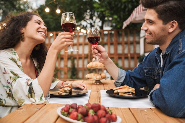 Ritratto di coppia sorridente uomo e donna che beve vino rosso mentre mangiano panini insieme a un tavolo di legno all'aperto