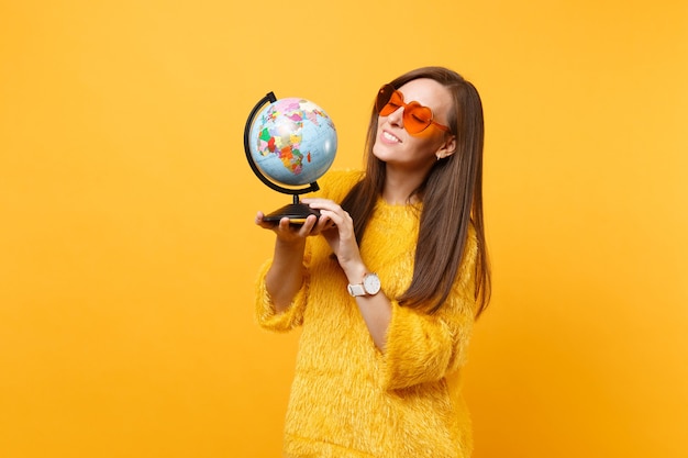 Ritratto di curiosa bella giovane donna in maglione di pelliccia occhiali cuore arancione guardando il globo del mondo isolato su sfondo giallo brillante. Persone sincere emozioni, concetto di stile di vita. Zona pubblicità.