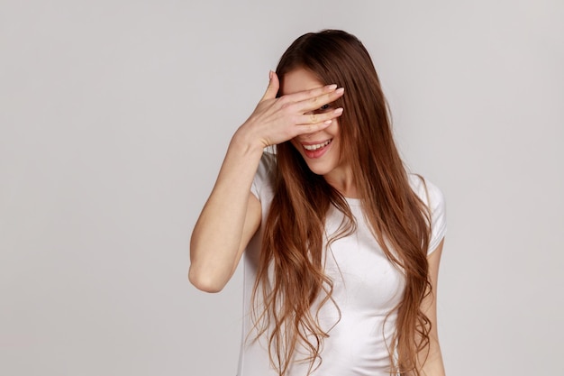 Ritratto di curiosa donna allegra con i capelli scuri in piedi che sbircia attraverso le dita alla telecamera spiando alla ricerca di un segreto che indossa una maglietta bianca Indoor studio girato isolato su sfondo grigio